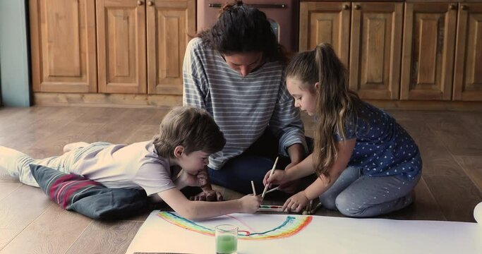 Mom with little son and 7s daughter sitting on warm floor holding brushes painting rainbow colorful picture with paints, enjoy time together, development of kids activity, modern comfort house concept
