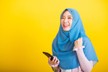 Asian Muslim Arab, Portrait of happy beautiful young woman Islam religious wear veil hijab funny smile she raise hand glad celebrating winning with smart mobile phone isolated on yellow background