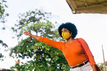 Young afro american woman using a face mask stopping a taxi on street, transport concept. 