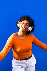 Smiling and cheerful afro woman dancing and listening to music with her wireless headphones, in front of a blue colored street wall, music technology concept.