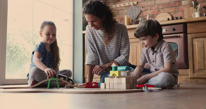 Pretty young mom with little 6s 7s children cute son and daughter sit on warm floor in kitchen engaged in game play wooden blocks and toy railroad. Funny leisure time during quarantine at home concept