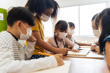 Young female teacher using an alcohol spray to disinfect student hands in classroom. Asian woman in face mask cleaning pupils' hands with hand sanitizer. School reopen after quarantine and lockdown. - Powered by Adobe