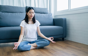 Beautiful Asian woman waking up in the morning practicing meditating exercise at home living room, routine healthy calm mind and body lifestyle spiritual mantra focus meditation lotus sitting position