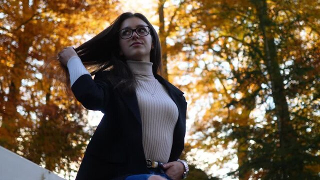 Slow Motion of a Young Woman on Fence of Public Park Under Trees in Fall Colors Touching Her Dark Hair, Low Angle