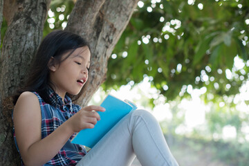  little asian girl reading a book under big tree. children and science. blurred background. learning the imagination and dreams of rural child. studying at home.