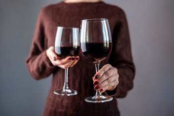 A woman in a brown sweater is holding two glasses of red wine and holding out one of them. Close up
