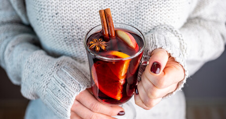 A woman in a white warm sweater is holding a transparent glass cup of mulled wine in her hand....