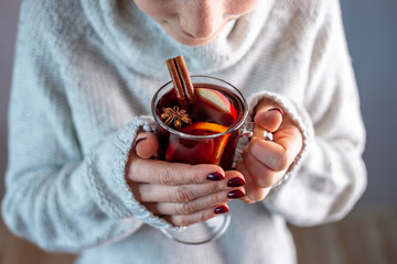 A woman in a white warm sweater is holding a transparent glass cup of mulled wine in her hand....