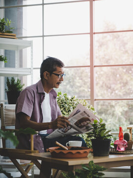 Asian Senoir Man Is Relaxing While Reading Newspaper In His Home House Plant Garden.