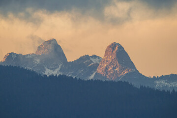peaks of the lions mountain range behind dense forest covered with few patches of snow under the beautiful orange light of setting sun over the North Vancouver