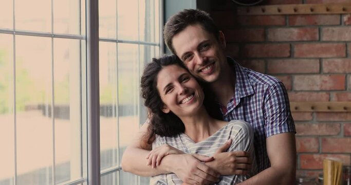 35s wife and husband embracing standing near panoramic window in modern kitchen, smiling look at camera feeling love and happiness at moving day at new first house, bank loan for young family concept