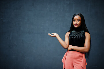 Fashionable african american woman in peach pants and black blouse pose against gray wall.