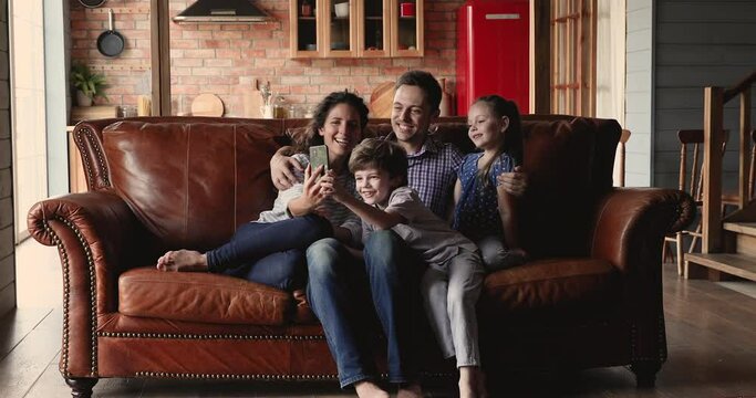 Family with little kids gathered in living room relaxing seated on couch using smartphone funny application, having fun with new app, take selfie photos, spend time on weekend with modern tech concept