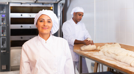 Portrait of smiling young hispanic female professional baker in white uniform standing in bakery during working day