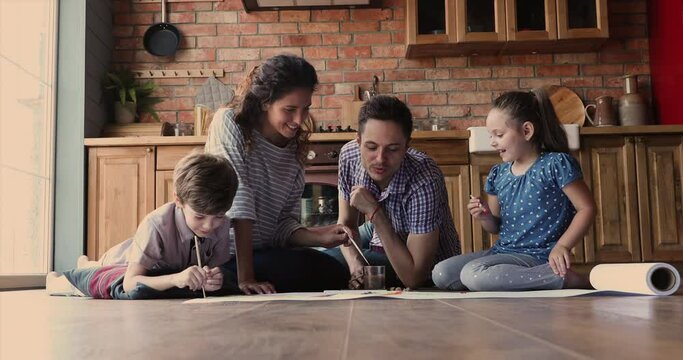 Young parents and preschool kids painting with paints on paper sheet seated on warm wooden laminate floor in cozy kitchen. Full family spend quarantine time with children at modern home, hobby concept