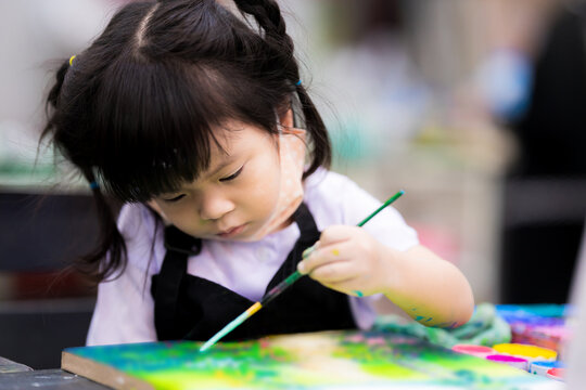 Asian girl paint art on canvas. Happy children are learning art. Cute children wear an apron during watercolor lessons. Child holds the paintbrush with his left hand. Adorable little kid 3 years old.