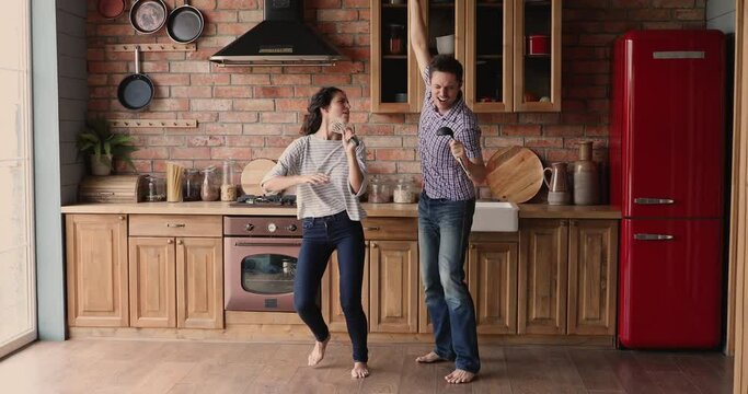 Cheerful girlfriend and boyfriend listen music dancing barefoot in kitchen sing song use kitchen utensil like microphone in microphones feels happy. Funny weekend activity, home karaoke, hobby concept
