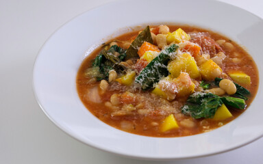 Ribollita or minestrone soup with beans, butternut squash, kale, bay leaf, and parmesan cheese served on the white shallow bowl. Diagonal view on white background.
