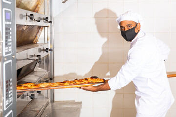 Baker in protective mask takes out an appetizing hot long bread called coca from the oven