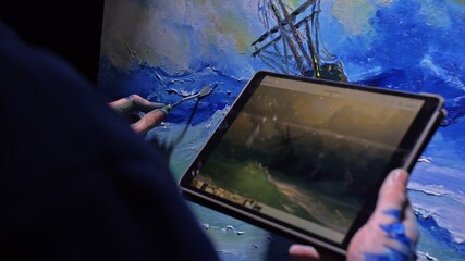 Artist copyist paint seascape with ship in ocean. Craftsman decorator draw as boat sail on blue sea with acrylic oil color. Draw finger, brush, knife palette. Indoor. In hands of tablet computer pc.