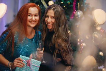 Obraz na płótnie Canvas Two Graceful women rejoices with a gift box near a Christmas tree. Women laughs, smiles, poses. Special vintage noise and grain filter, blurry lights.