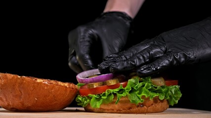 Craft burger is cooking on black background in black food gloves. Consist: sauce, lettuce, tomato,...