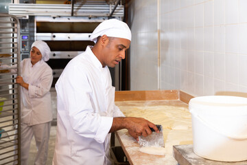 Skilled Hispanic bakery worker portioning dough with scraper and weighing pieces