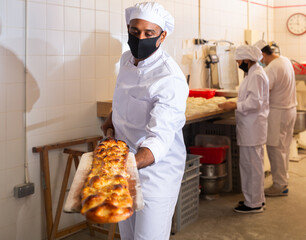 Baker in protective mask takes out an appetizing hot long bread called coca from the oven