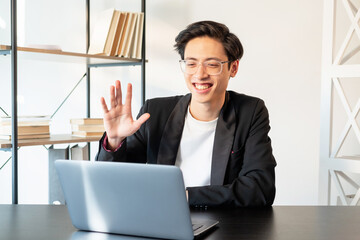 Video conference. Virtual meeting. Online communication. Cheerful asian business man in glasses greeting team waving hand on laptop camera in light home office.