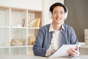 Distance learning. Online education. Virtual class. Cheerful smart asian male student in white wireless headphones glasses listening lesson with copybook looking at camera at light workplace.