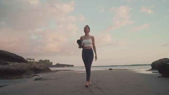 Full tracking low angle of sun-tanned toned Caucasian girl with hair up wearing leggings and cropped tank top holding rolled up yoga mat walking by sandy ocean shore on tropical island at sunset