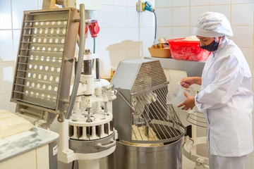 Tuinposter Young female bakery worker controlling dough kneading on professional equipment © JackF