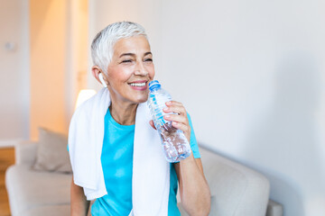 Elderly woman sitting on the sofa, exhausted after daily training. Senior woman taking a break while exercising at home. Athletic mature woman holding bottle of water,Having a towel around her neck