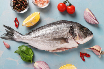 Fresh ready to cook raw fish dorado with ingredients and seasonings like basil, lemon, salt, pepper, cherry tomatoes and garlic on light blue background, top view