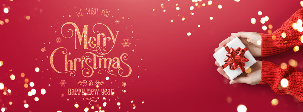 Female hands holding Christmas gift box with red bow on red background with bokeh, light and Merry Christmas and Happy New Year text. Xmas greeting card, wide holiday banner. Flat lay, top view
