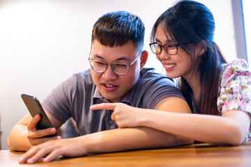 Happy asian couple in coffee shop using smartphone together, smiling young couple embracing while looking at smartphone.
