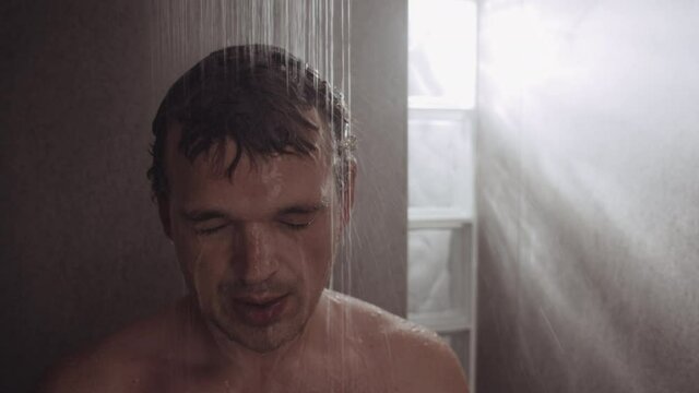 Close-up shot a man standing under the shower and getting relaxed with cool water, then he coming up to the camera with relieved look