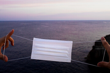 Two hands holding a white, surgical mask that stands out on the sea at sunset