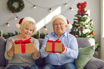 Obraz na płótnie Canvas Happy grandma and grandma in Santa hats sitting on sofa holding Christmas presents for grandkids