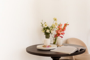 Bouquets of fresh flowers and open magazine placed on table near clean dishware against beige wall...
