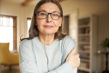 Horizontal portrait of senior gray haired European female in eyeglasses posing indoors, looking at...