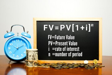 A picture of alarm clock, fake cash, coins and blackboard with time value of money formula on table. 1 dollar in future is not equivalent to today value. 