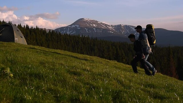 Couple of tourists with backpacks approach the tent town in the mountains and enjoy the beautiful view. Tents stand on a hill against a wonderful spruce forest and powerful mountains under a blue sky.