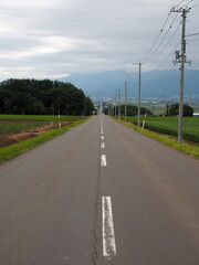 Fototapeta na wymiar A long straight road with mountains in the background on cloudy summer day in Hokkaido, northern Japan