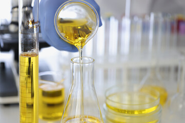 Hand in rubber glove pours yellow liquid into flask in chemical laboratory closeup. Pharmacological preparations production concept.