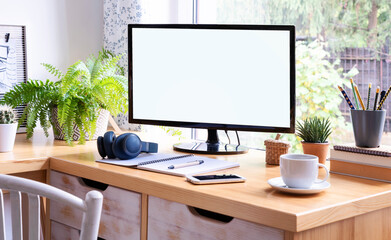 Wooden office desk with empty screen computer with notebook, pen, cans, mobile and plant near window. Workplace interior during home office. Mock up.