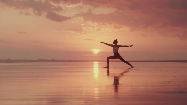 Wide shot of slim unrecognizable female silhouette standing with legs separated into wide stance. Girl stretching on sandy ocean shore, beautiful sky on background, sun setting over horizon