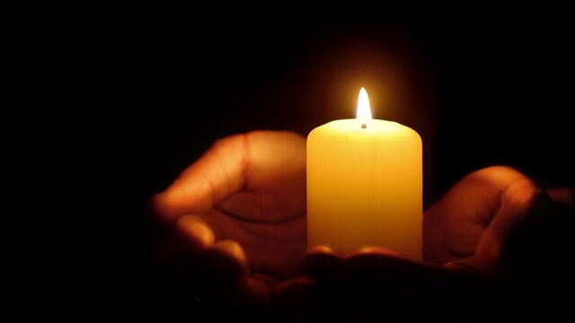 Burning candle resting on steady female palms, glowing serene light in the dark, low-key shot with black background and copy space 
