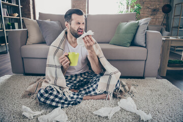 Obraz premium Full length photo of sick ill guy sit carpet hold hot tea beverage mug sneeze napkin cover plaid checkered blanket wear nightwear sleepwear in house indoors