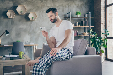 Photo of focused man sit divan use smartphone search read coronavirus information wear checkered plaid pants trousers white t-shirt in house indoors